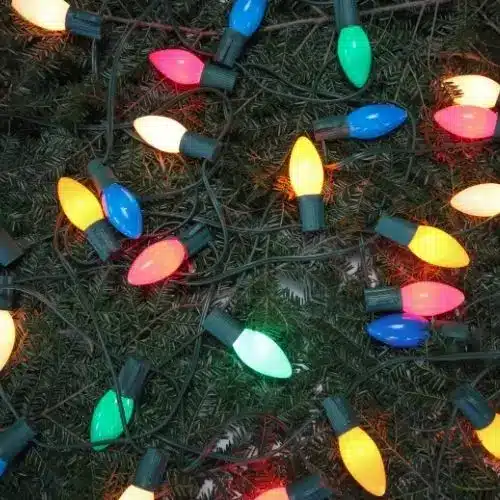 A dense cluster of classic, multi-colored C7 or C9 Christmas light bulbs tangled on a bed of dark green pine tree branches, featuring red, blue, green, yellow, and pink colors.